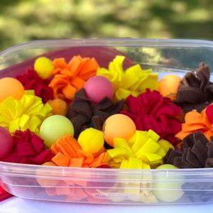 May include: A clear plastic container filled with colourful felt balls and pom-poms. The balls and pom-poms are in shades of red, orange, yellow, and brown.