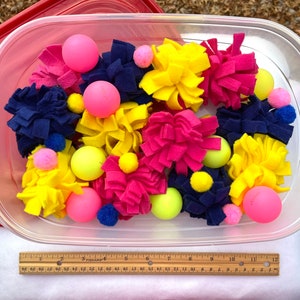 May include: A clear plastic container filled with colourful craft supplies. The container holds felt flowers in shades of pink, yellow, and navy blue, along with pink and yellow balls and small pom-poms. A ruler is visible at the bottom of the container.