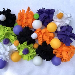May include: A collection of colourful felt balls and pom-poms in various shades of orange, green, purple, white, and black. The balls are arranged in a cluster on a white surface.