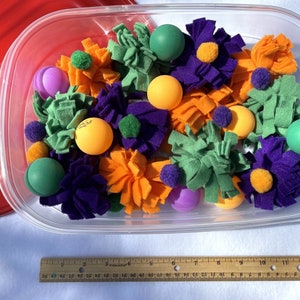 May include: A clear plastic container filled with a variety of colourful felt toys. The toys include green, orange, purple, and yellow felt flowers, as well as green and orange felt balls.
