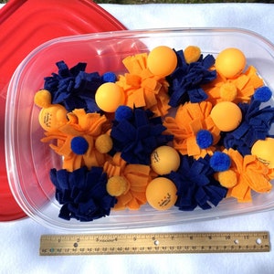 May include: A clear plastic container filled with blue and orange felt toys. The toys are shaped like pom-poms and have small orange balls in the centre.