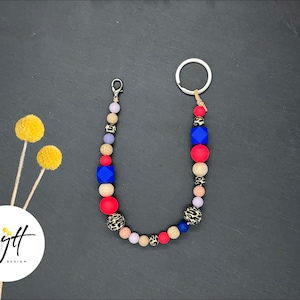 Può includere: Un portachiavi con perline con una varietà di perline colorate, tra cui rosse, blu e con stampa leopardata. Il portachiavi ha una chiusura argentata e un portachiavi. Il design include il testo "Lytt Design" in un cerchio bianco.