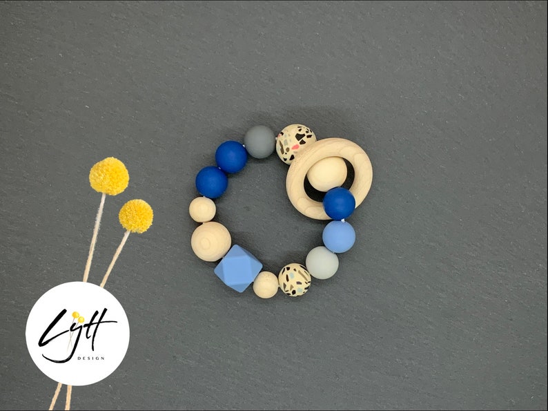K&ouml;nnte beinhalten: Ein Baby-Bei&szlig;ring mit einem Holzring und einer Vielzahl farbiger Perlen. Die Perlen sind in Blau-, Grau-, Creme- und Sprenkelmustern gehalten. Das Design umfasst eine geometrische blaue Form und einen Holzring.