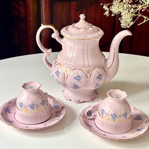 May include: A light pink porcelain tea set consisting of a teapot with a lid and two inverted teacups on matching saucers. The set features gold-trimmed scalloped edges and a delicate blue floral pattern within arched panels on each piece.