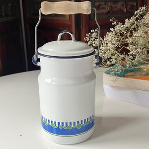 May include: A white enamel milk churn with a lid and a metal handle featuring a light wood grip. The base of the container is decorated with a horizontal blue band containing a repeating pattern of small green circles and thin white lines.