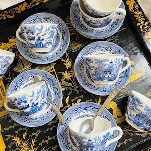 Può includere: Una collezione di tazze e piattini in porcellana blu e bianca con il classico motivo Willow. Le tazze sono bianche all'interno e hanno un manico. Cucchiaini d'argento sono appoggiati in alcune tazze. Il set è disposto su un vassoio nero con dettagli dorati.