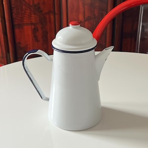 May include: A white enamel coffee pot with a tapered body, a matching lid featuring a small red knob, a dark blue rim, and a dark blue handle. A long, curved red spout extends from the side of the pot, which has a smooth, glossy finish throughout.