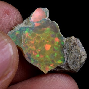 May include: A rough piece of opal with a green, orange, and red iridescent sheen. The opal is set in a matrix of grey rock.