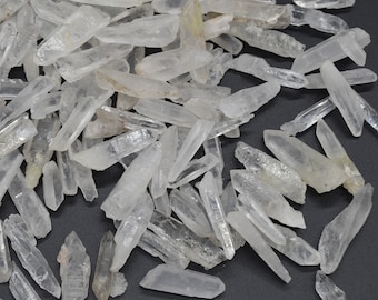 May include: A collection of clear quartz crystal points, also known as rock crystals, in various sizes and shapes. The crystals are scattered on a dark gray surface.