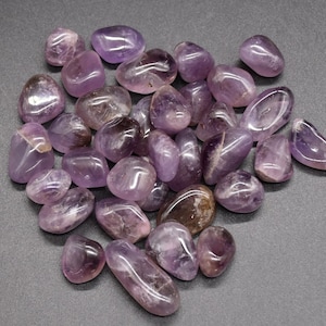 May include: A selection of polished amethyst stones in shades of purple, from pale lavender to deep violet. The stones are smooth and rounded, with natural colour variations and some translucent patches. They are scattered on a grey surface.