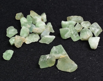 Pode incluir: Uma coleção de cristais de fluorita verde, alguns com um leve tom amarelo, sobre um fundo preto. Os cristais são ásperos e têm uma variedade de formas e tamanhos.