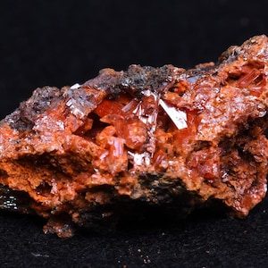 Könnte beinhalten: Ein Cluster aus leuchtend orangefarbenen Kristallen, eingebettet in einem dunkelbraunen Gestein. Die Kristalle sind scharf und haben einen glasartigen Glanz.