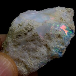 May include: A rough, white opal with a green and pink iridescent sheen. The opal is embedded in a matrix of gray rock.