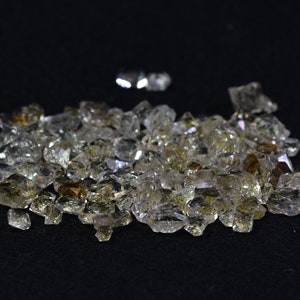 May include: A pile of small, clear quartz crystals with a slight golden hue. The crystals are scattered on a black surface.