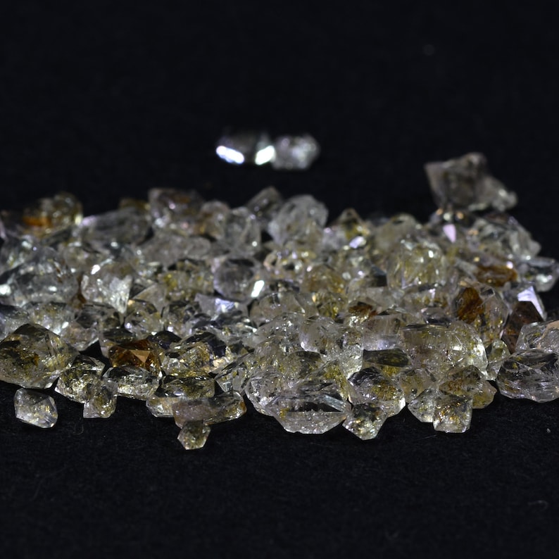 May include: A pile of small, clear quartz crystals with a slight golden hue. The crystals are scattered on a black surface.