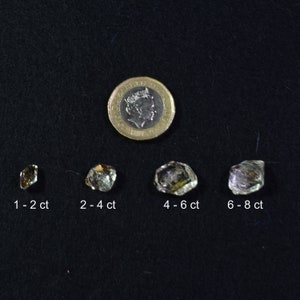 May include: Four clear, faceted crystals of varying sizes, displayed on a black surface. The crystals are labeled with their approximate carat weight: 1-2 ct, 2-4 ct, 4-6 ct, and 6-8 ct. A British one pound coin is included for scale.