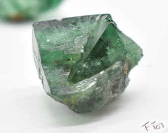 May include: A green fluorite crystal with a sharp, geometric shape. The crystal has a translucent appearance and is partially covered in a layer of white mineral.