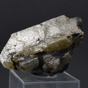 May include: A rough mineral specimen with a layered, crystalline structure. The stone has a pale green and silver colour, with dark grey and black inclusions. The specimen is displayed on a clear, angled stand against a dark background.