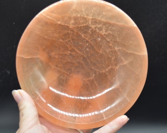 May include: A round, light orange, translucent, polished, decorative dish made of natural stone. The dish has a slightly raised rim and a smooth, polished surface.