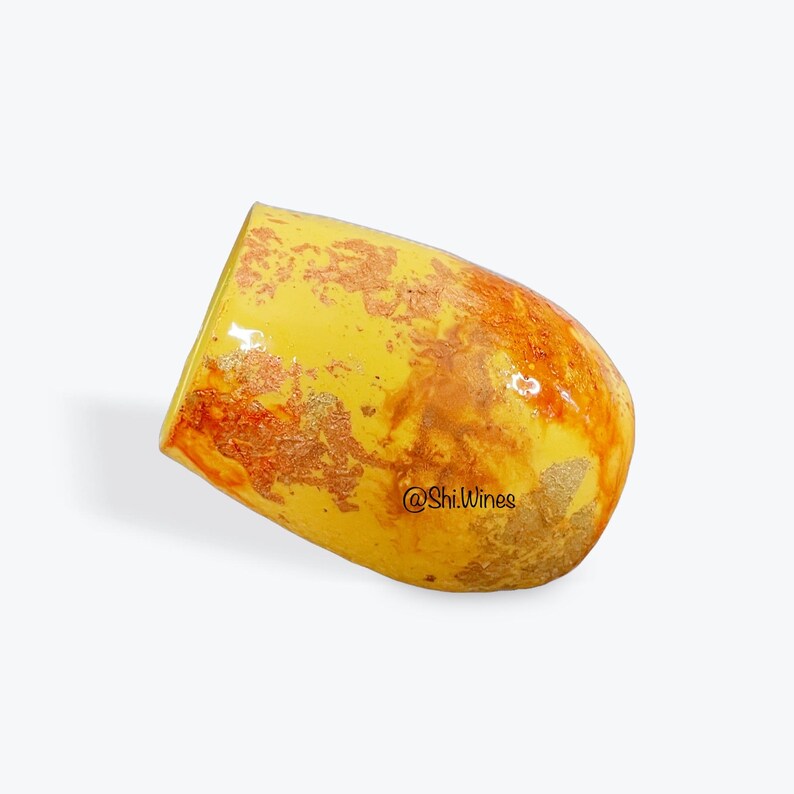 May include: A yellow stemless wine glass with a textured surface and gold and orange accents. The glass has a unique shape and is tilted slightly to the side. The text "@Shi.Wines" is visible on the side of the glass.
