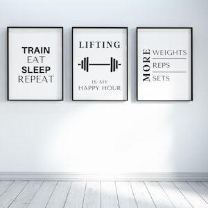 May include: Three framed prints with black frames and white backgrounds. The prints feature black text related to fitness: "TRAIN EAT SLEEP REPEAT", "LIFTING IS MY HAPPY HOUR" with a barbell graphic, and "MORE WEIGHTS REPS SETS".