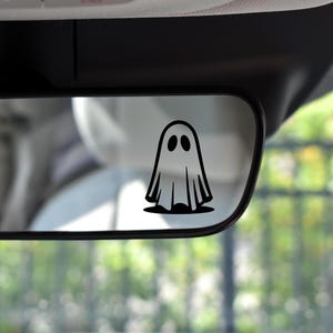 May include: A black ghost decal is affixed to a car's rearview mirror. The ghost has a simple, cartoonish design with two oval eyes and a flowing sheet-like body. The background shows a blurred view of the car's interior and exterior.