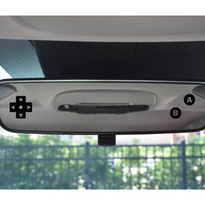 May include: A car's rearview mirror with a black directional pad graphic on the left side and the letters "A" and "B" in black circles on the right. The mirror is mounted on the windshield, with a view of a fence and greenery in the background.