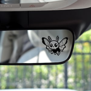 May include: A black and white sticker of a cartoon moth with a skull head and large eyes, attached to a rearview mirror in a car.