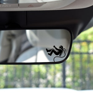 May include: A rearview mirror with a black astronaut sticker on it. The astronaut is floating in space with a tether.