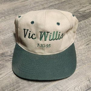 May include: A beige and green baseball cap with the text "Vic Willis 7-30-95" embroidered on the front.
