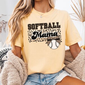 May include: Butter-colored t-shirt with the text "SOFTBALL Mama" in black and leopard print. A softball with a leopard print bow is featured below the text. The shirt is a short-sleeved crew neck.