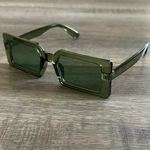 May include: Green square-shaped sunglasses with a dark green lens. The sunglasses are made of plastic and have a modern design.