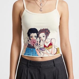 Op de afbeelding: Crèmekleurige tanktop met een kleurrijke anime-achtige afbeelding van twee vrouwen die zich opmaken. De top heeft dunne bandjes en wordt gecombineerd met een donkergrijze geplooide broek. Een gelaagde ketting en armband zijn ook zichtbaar.