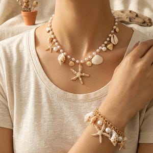 Op de afbeelding: Een goudkleurige ketting en armband set met schelp- en zeesterbedels. De ketting heeft imitatieparels en schelpenhangers. De armband heeft vergelijkbare bedels. De sieraden zijn ontworpen in een strand- of nautische thema.