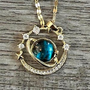 May include: Gold necklace with a blue and green galaxy-like pendant. The pendant is surrounded by a crescent moon and stars, all set with clear rhinestones.