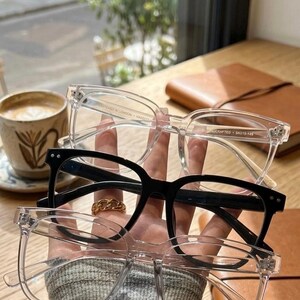 May include: A hand holds three pairs of eyeglasses: two clear-framed and one black-framed. The clear frames have a rectangular shape, while the black pair has a square shape. A gold chain ring is visible on a finger. A cup of coffee sits in the background.
