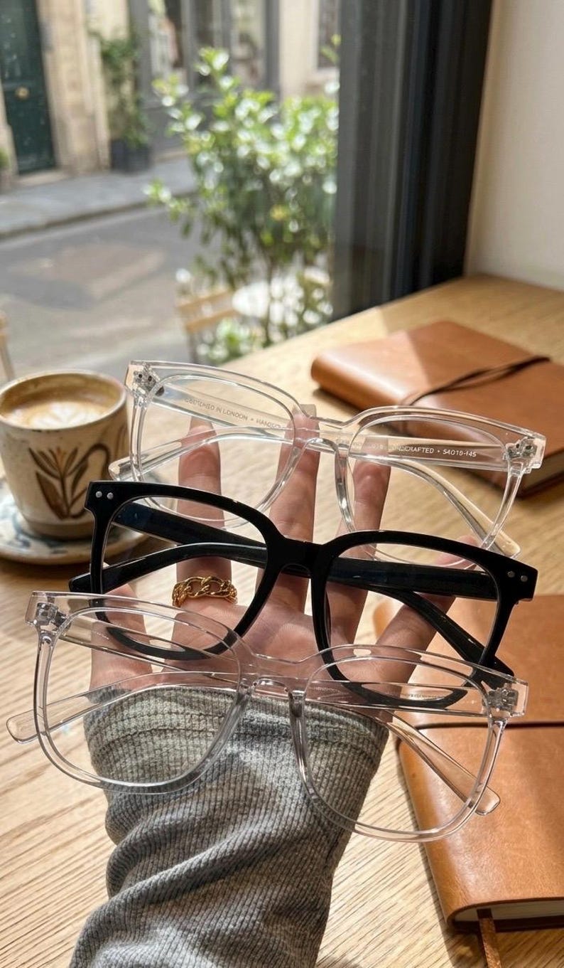 May include: A hand holds three pairs of eyeglasses: two clear-framed and one black-framed. The clear frames have a rectangular shape, while the black pair has a square shape. A gold chain ring is visible on a finger. A cup of coffee sits in the background.