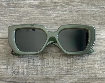 Könnte beinhalten: Eine Sonnenbrille mit quadratischen Gläsern in Dunkelgrün und einem dicken Rahmen.