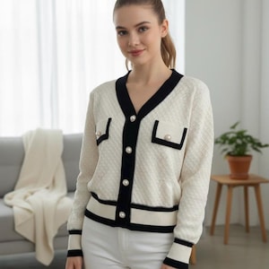 May include: Cream-colored cardigan with black trim and pearl-like buttons. The V-neck sweater features two small pockets and is worn with white pants. The cardigan has a textured pattern and is a classic style.