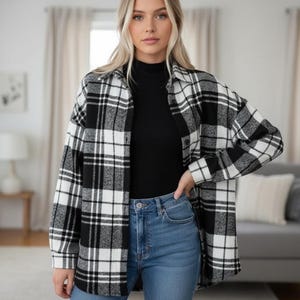 May include: A black and white plaid flannel shirt. The shirt is open, revealing a black turtleneck and blue jeans. The flannel has a classic pattern with large squares of black and white. The sleeves are long and the shirt appears to be made of a soft material.