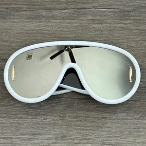 Puede incluir: Gafas de sol aviador blancas con lentes espejados. Las gafas tienen una montura blanca gruesa y una forma redondeada.