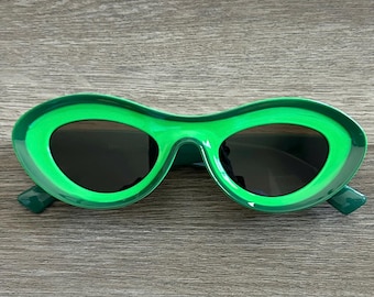May include: Green oval-shaped sunglasses with a thick green frame and dark lenses.