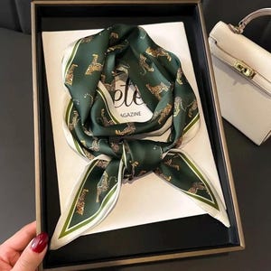 May include: A dark green silk scarf with a leopard print design. The scarf has a white border with a green stripe. The scarf is knotted and lies on a white surface with the word "ete" visible. A white purse is in the background.