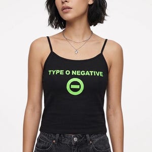 以下が含まれることがあります： 明るい緑色の「TYPE O NEGATIVE」の文字と、水平線が入った緑色の円が特徴の、細いストラップが付いた黒いタンクトップ。濃い色のデニムジーンズと合わせています。