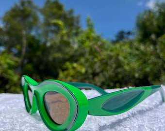 May include: Bright green sunglasses with a unique, rounded frame design. The lenses are a light brown color, and the sunglasses are set against a backdrop of green foliage and a blue sky. The sunglasses are a stylish accessory.