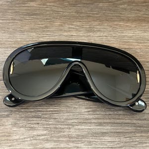 Op de afbeelding: Zwarte oversized zonnebril met een afgerond, futuristisch montuur. De glazen zijn donker en het montuur heeft een gouden accent.