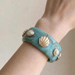 Peut inclure: Un large bracelet jonc en résine turquoise marbrée. Le bracelet est orné d'une alternance de breloques en forme de coquillage en métal doré et de perles blanches artificielles rondes serties dans des chatons en métal doré sur la surface.