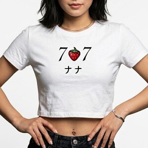 May include: White crop top with a graphic design featuring the numbers "7 7", a red strawberry, and Japanese characters. The shirt is a casual style.
