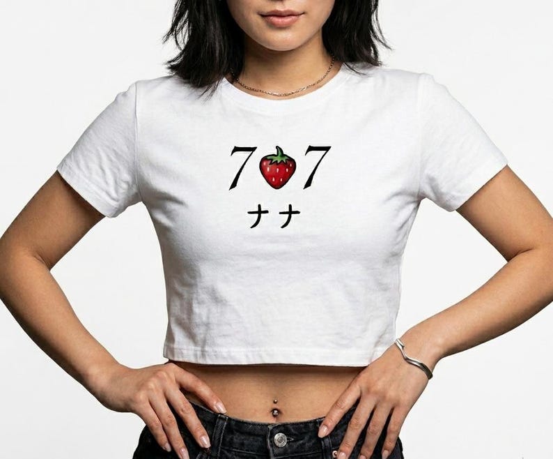 May include: White crop top with a graphic design featuring the numbers "7 7", a red strawberry, and Japanese characters. The shirt is a casual style.