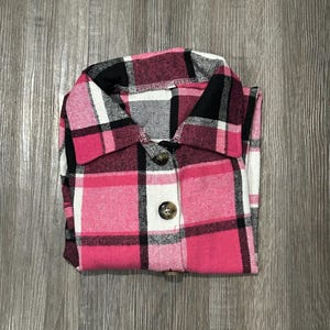 Op de afbeelding: Een roze, zwart en wit geruit flanellen overhemd met bruine knopen.